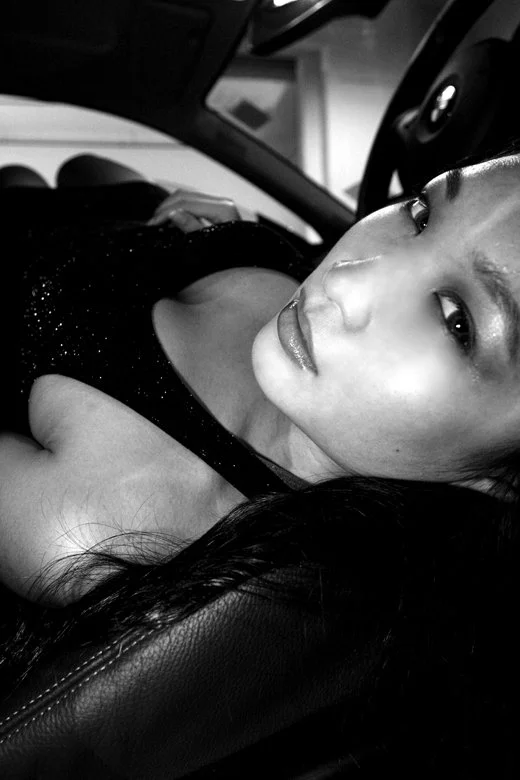 A woman with long dark hair and makeup, sitting in a car, looking at the camera with an intense expression.