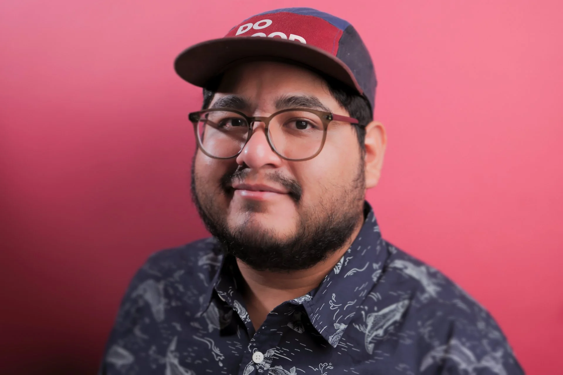 A man with glasses, a beard, and a mustache wearing a cap and a patterned shirt, smiling at the camera against a pink background.