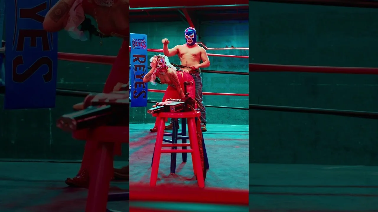 Two luchadores wearing masks, one is shirtless and flexing, the other is sitting on a red stool with a mask and red outfit, inside a wrestling ring with red ropes and a green wall background.