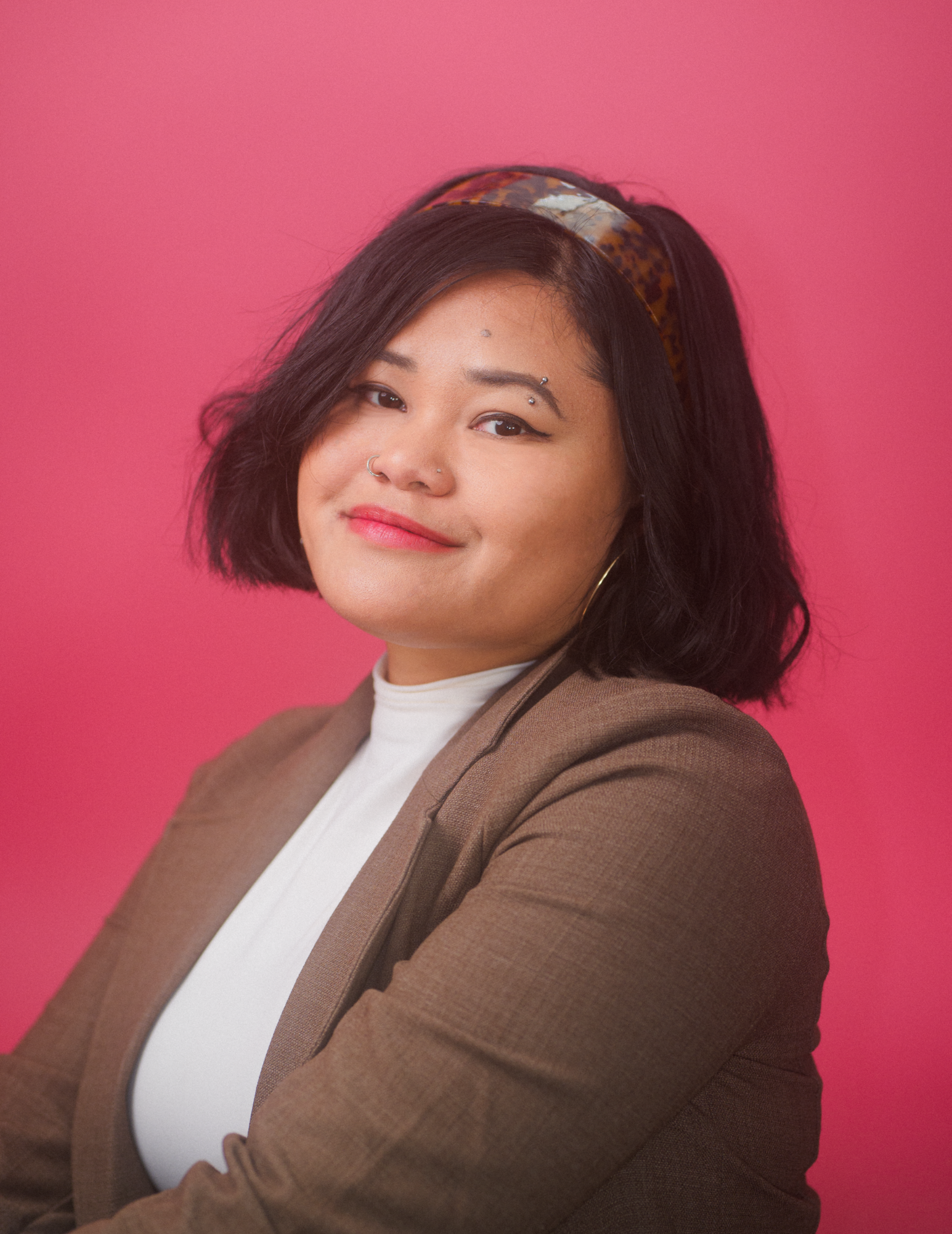 A woman with dark hair styled in a bob, wearing a brown blazer over a white top, with a patterned headband and multiple piercings on her face, standing against a pink background.