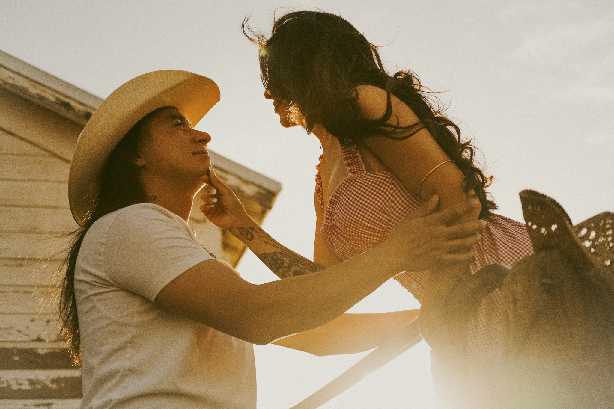 Two women sharing an intimate moment outdoors during sunset, one with curly hair and the other with straight hair and a hat.