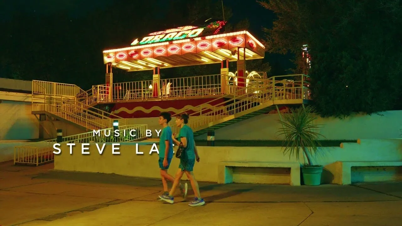 Nighttime scene at an amusement park with a brightly lit ride, two people walking, and text indicating music by Steve La.