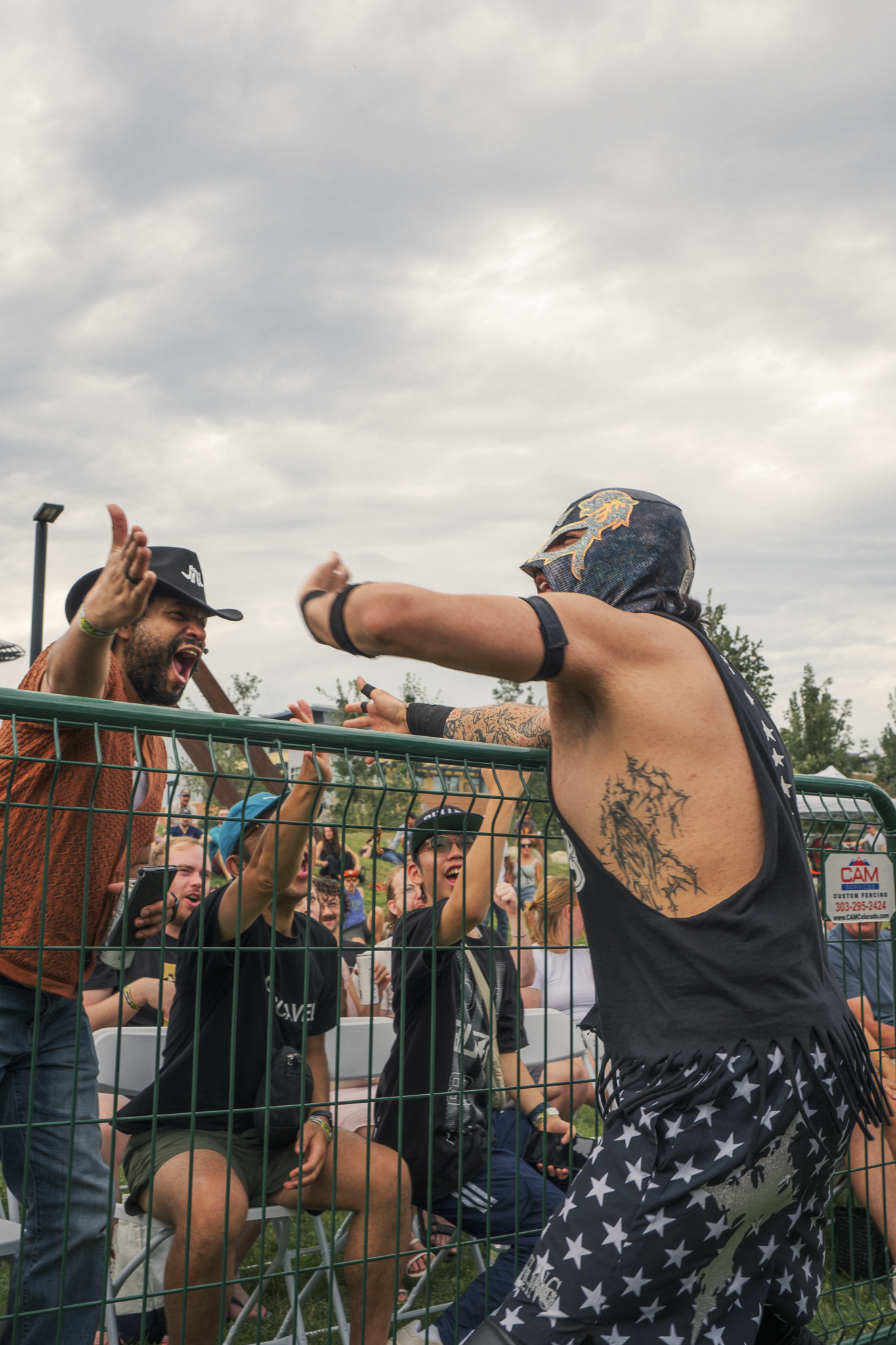 A group of people at an outdoor event, interacting excitedly through a metal fence, with a man wearing a lucha libre mask and star-patterned shorts at the forefront.