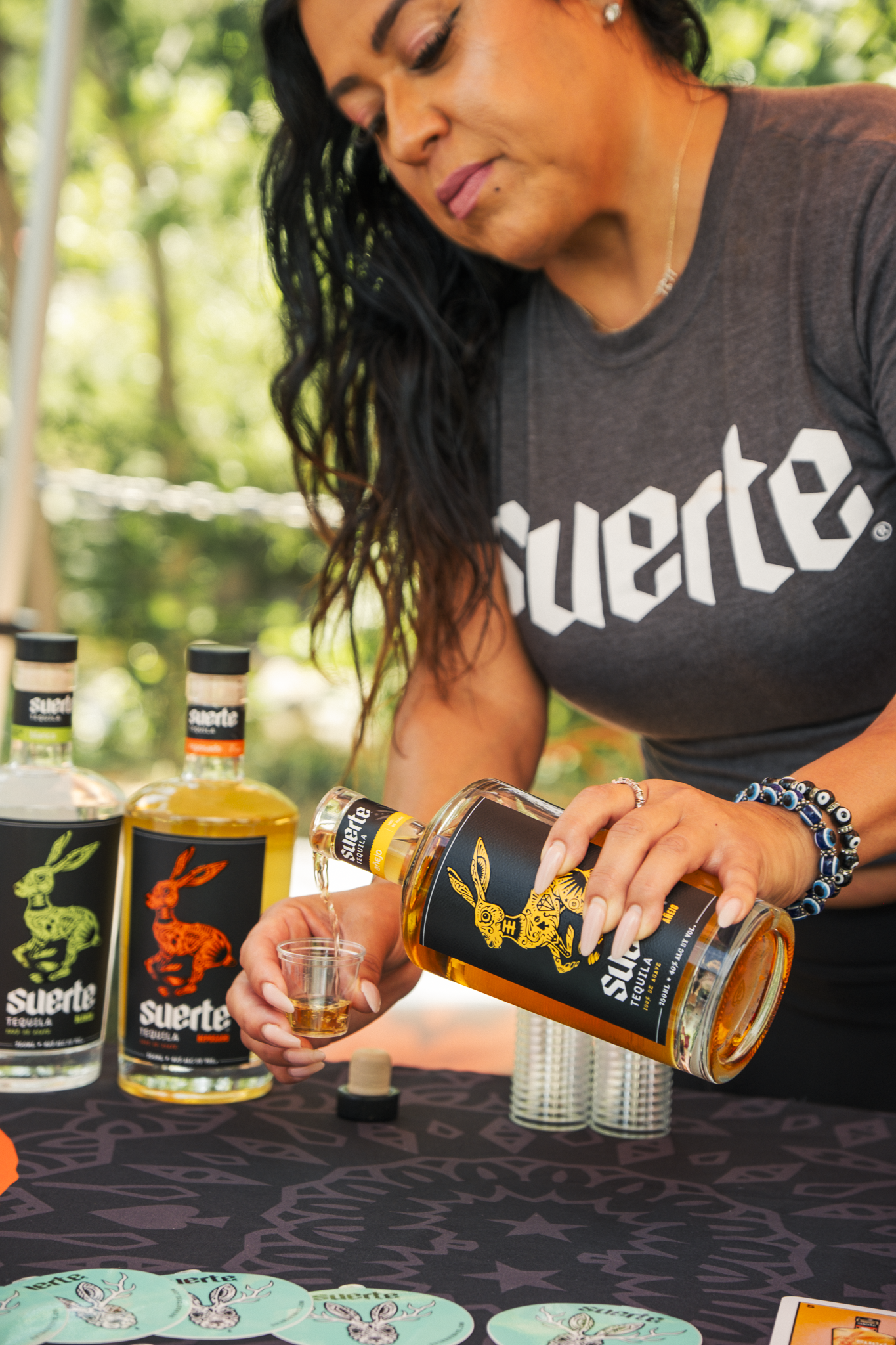 A woman with black curly hair pouring tequila from a bottle into a small shot glass. She is wearing a gray T-shirt with the word 'suerte' and a rabbit logo, and several bracelets. There are multiple bottles of 'suerte' tequila on the table, along wit