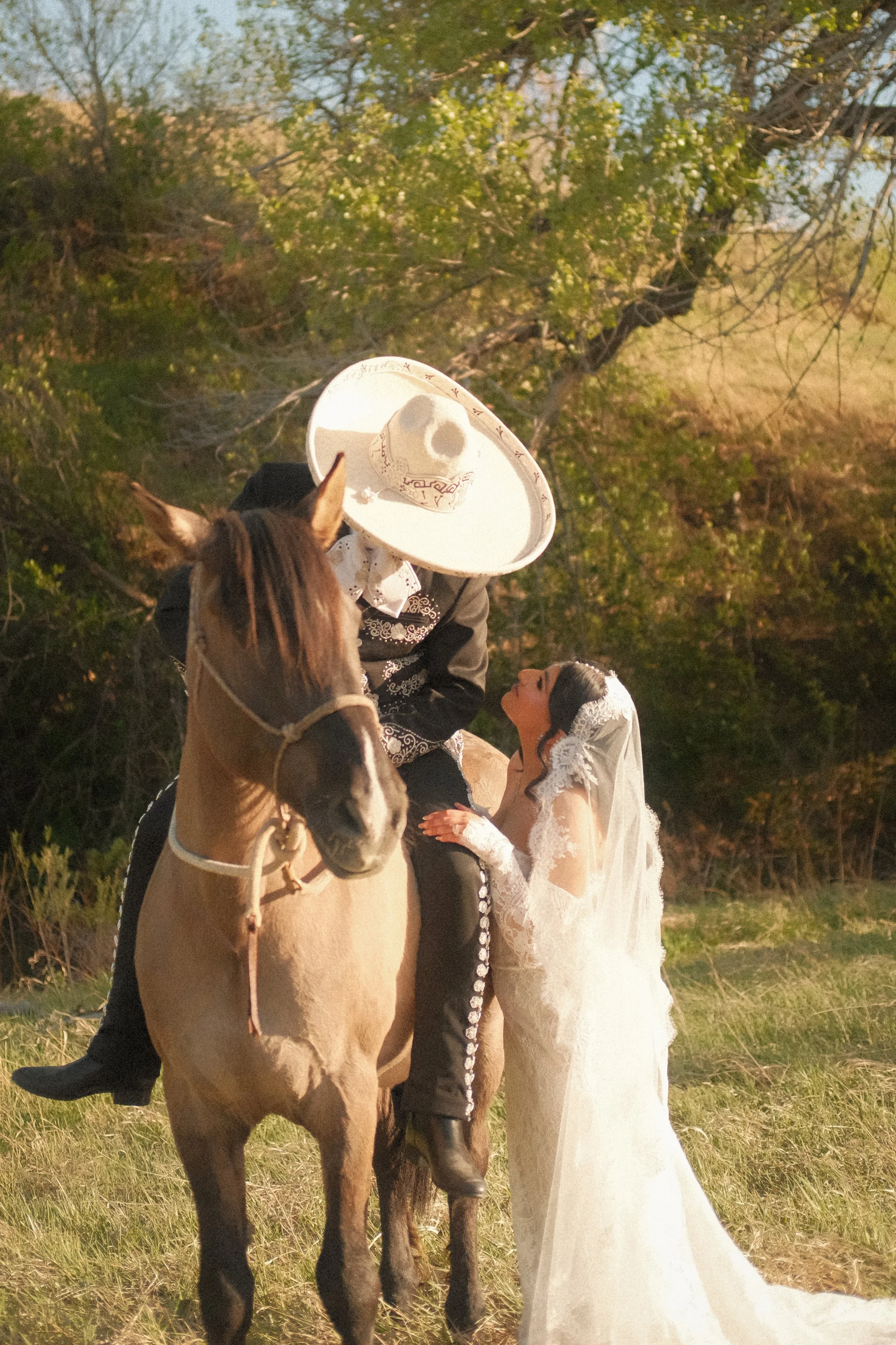 A woman dressed in a white wedding gown and veil standing next to a horse with a rider wearing traditional Mexican charro attire, including a large white sombrero, in an outdoor setting with green trees and grass.