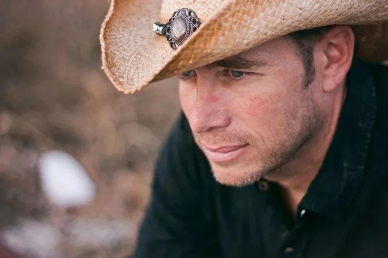 A man wearing a cowboy hat with a decorative pin, looking thoughtfully downward. He has facial hair and is dressed in a dark shirt, outdoors.