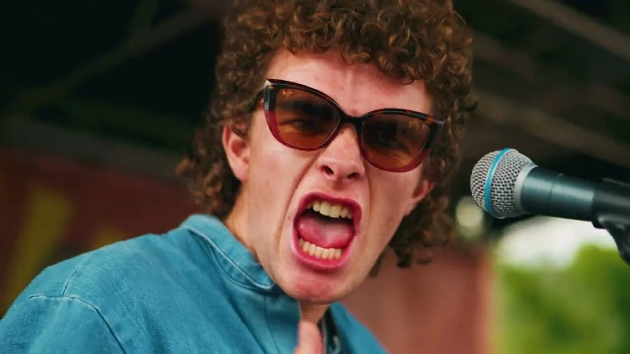 Young man with curly hair and sunglasses yelling into a microphone.