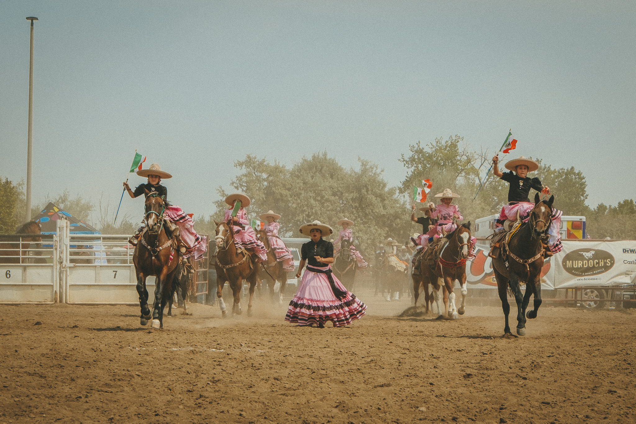 Group of women dressed in traditional Mexican attire riding horses at a rodeo or parade, waving Mexican flags.