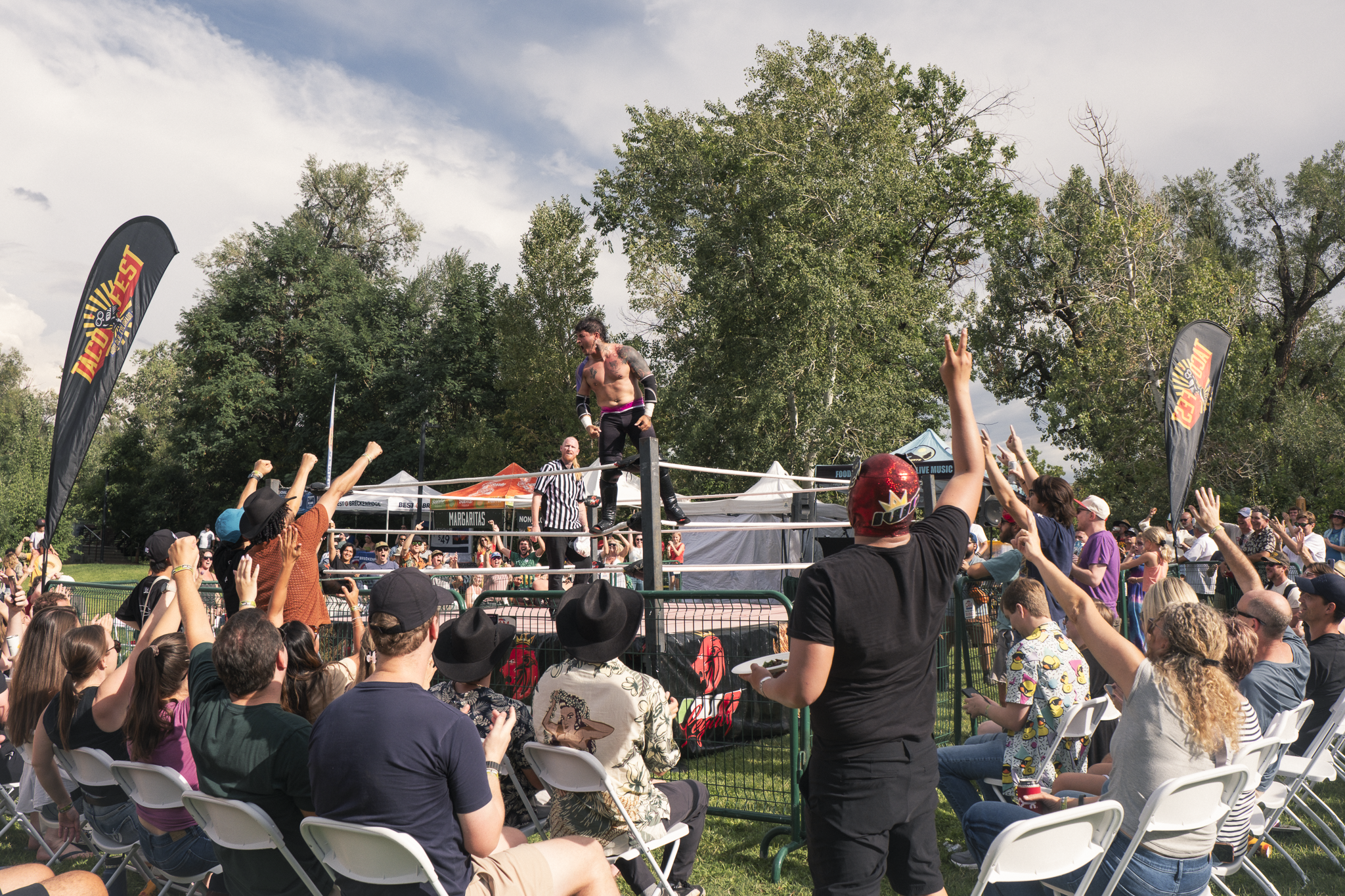 Crowd watching a pro wrestling match outdoors with a male wrestler standing on the ring, raising his fists, surrounded by trees and banners.