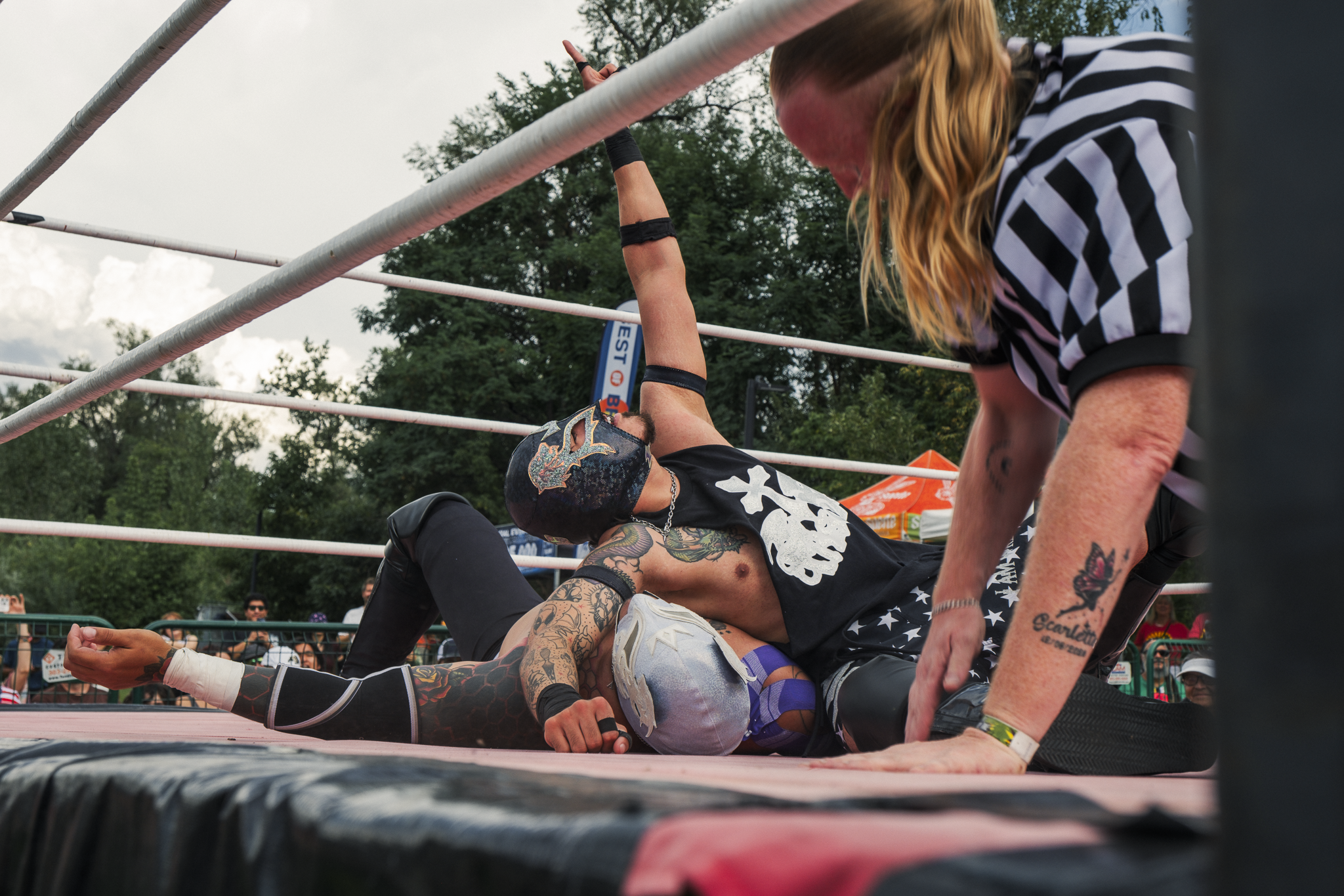 A wrestling match outdoors with one wrestler lying on the mat and another wrestler pinning him down, while a female referee in a black and white striped shirt looks on. Spectators are visible in the background.