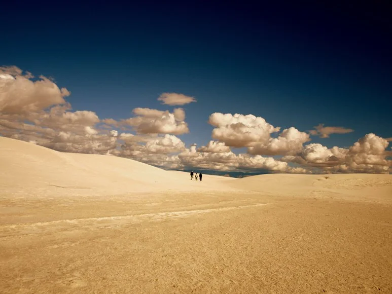 Three people walking across a vast, sandy, desert-like landscape with rolling dunes under a partly cloudy sky.