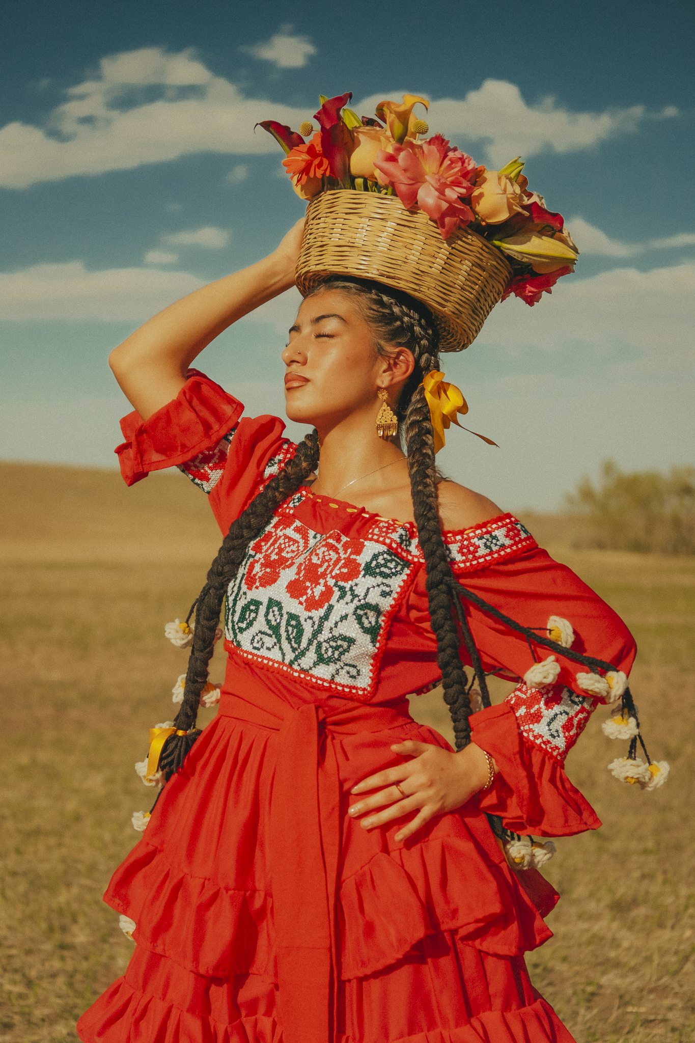 A woman dressed in traditional Ukrainian clothing carrying a basket of flowers on her head with her eyes closed in a field under a blue sky with clouds.