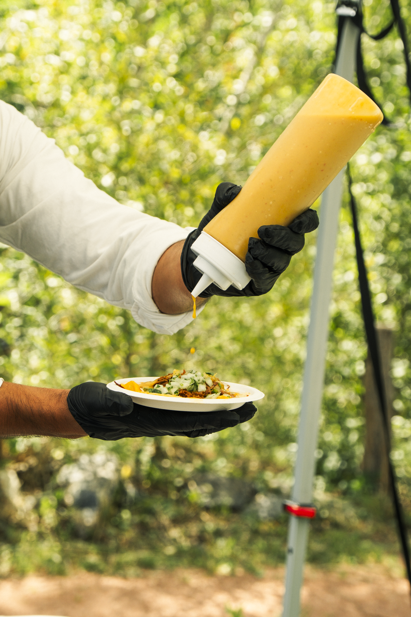Person holding a plate of food while a second person sprays sauce onto it outdoors with trees in the background.