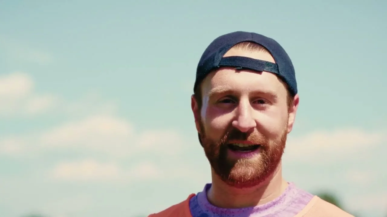 A man with a beard wearing a backward navy blue cap and a pastel purple shirt outdoors against a partly cloudy sky.
