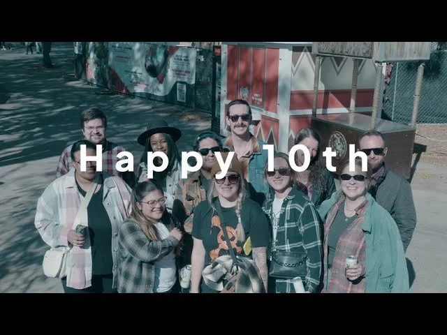 Group of nine people smiling outdoors at a celebration or festival, with the words "Happy 10th" overlayed on top.