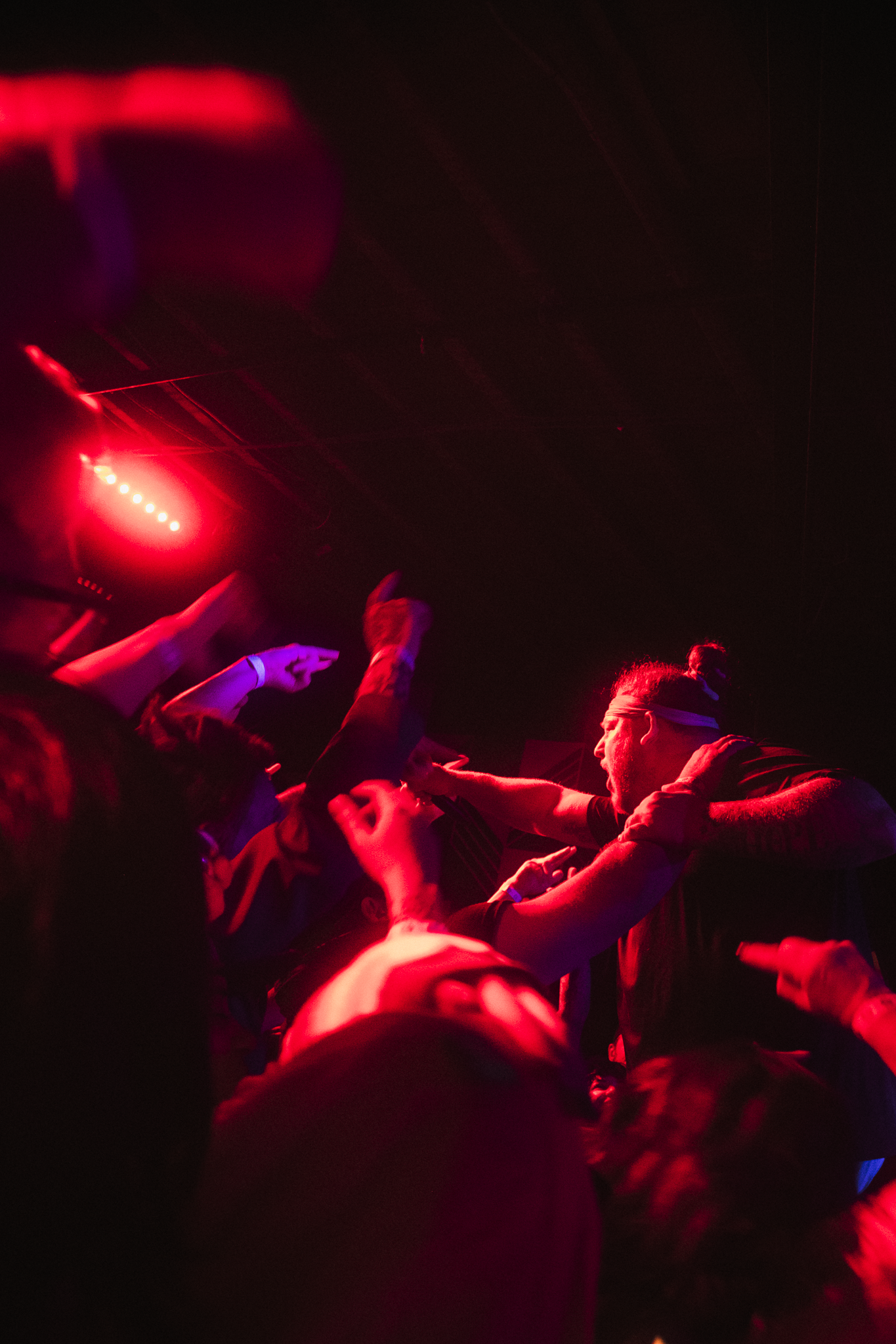 People in a dark, crowded space illuminated by red light, reaching out and holding onto each other in an energetic scene.