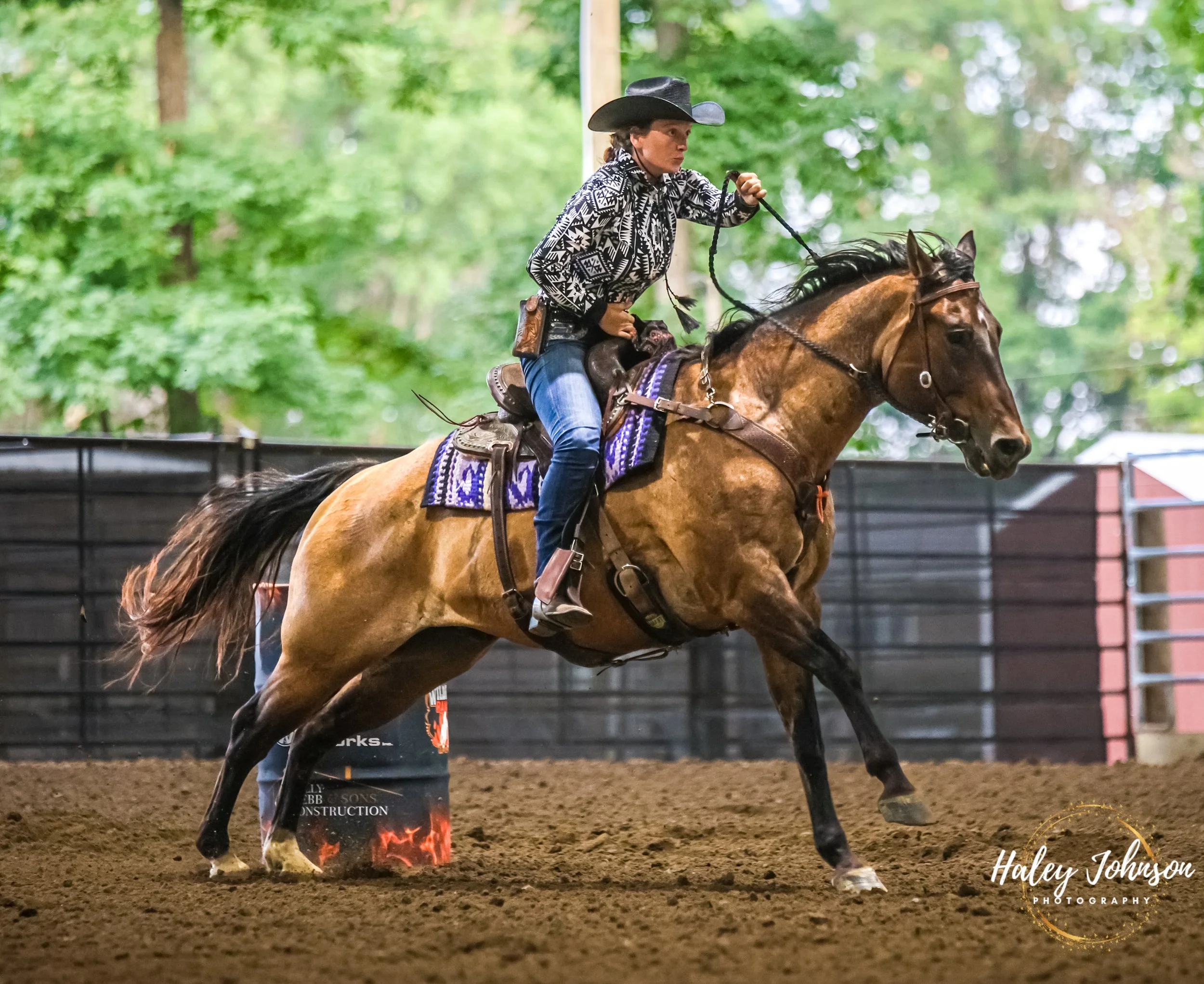 A woman riding a galloping horse through the barrel pattern in a covered arena, wearing a black cowboy hat and a patterned black and white shirt.