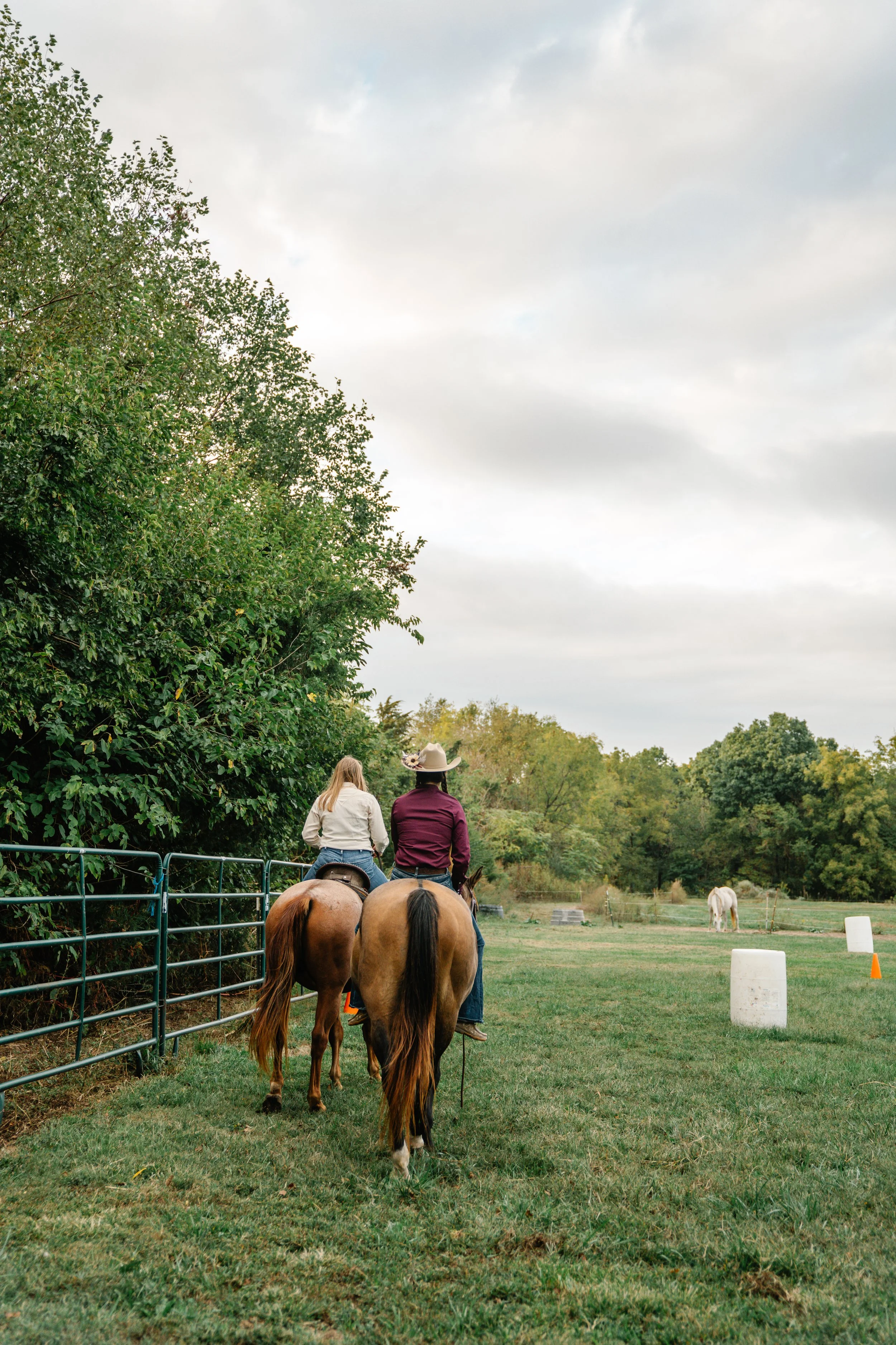 Two people riding horses along a farm fence, with trees on one side and a field with another horse in the background, under a cloudy sky.
