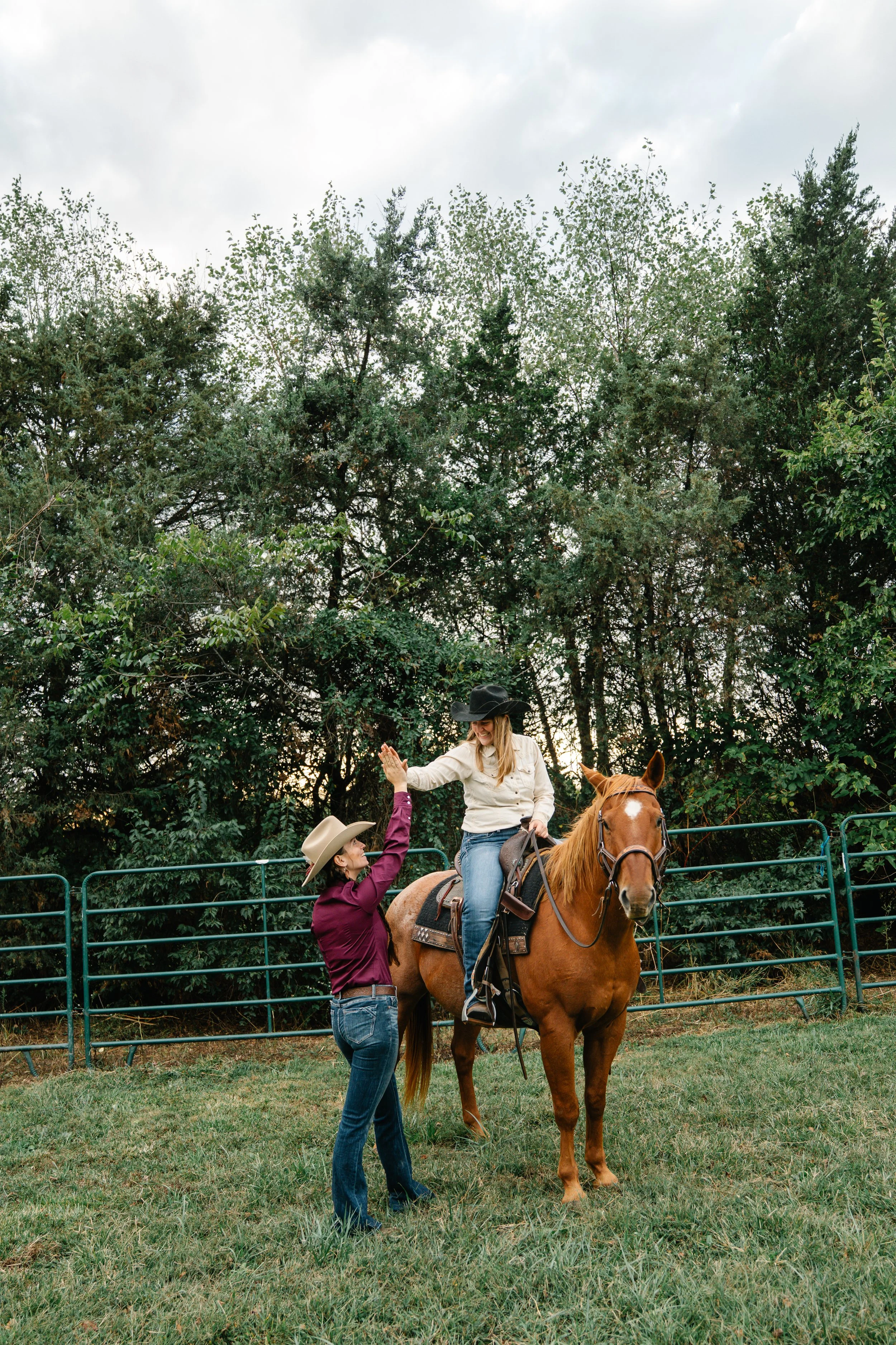 A woman on horseback giving a high five to a woman in a cowboy hat standing outside in a fenced area with tall trees and overcast sky.