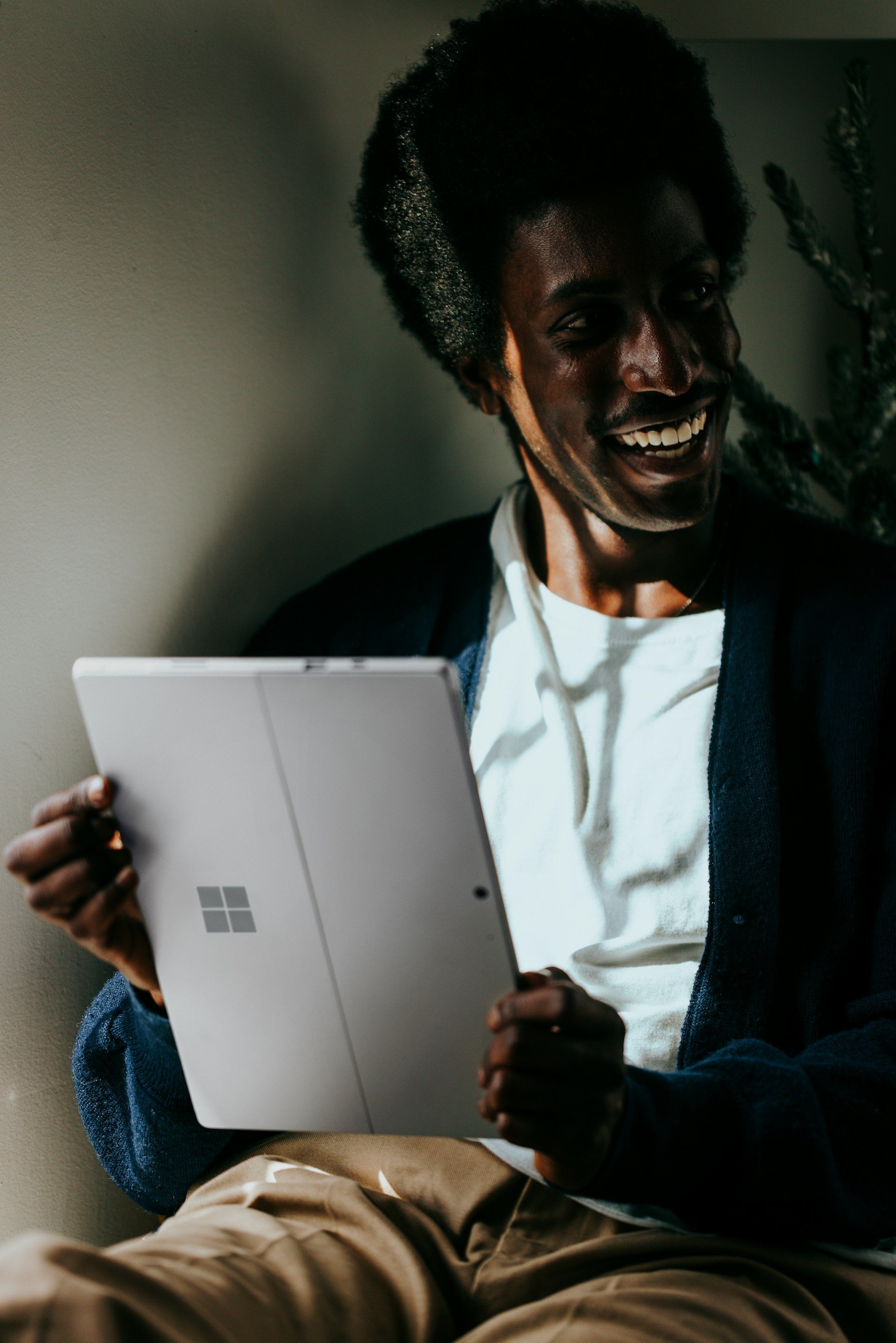 Young man with dark skin and afro hair smiling and holding a white tablet device, sitting against a plain wall with a plant in the background.