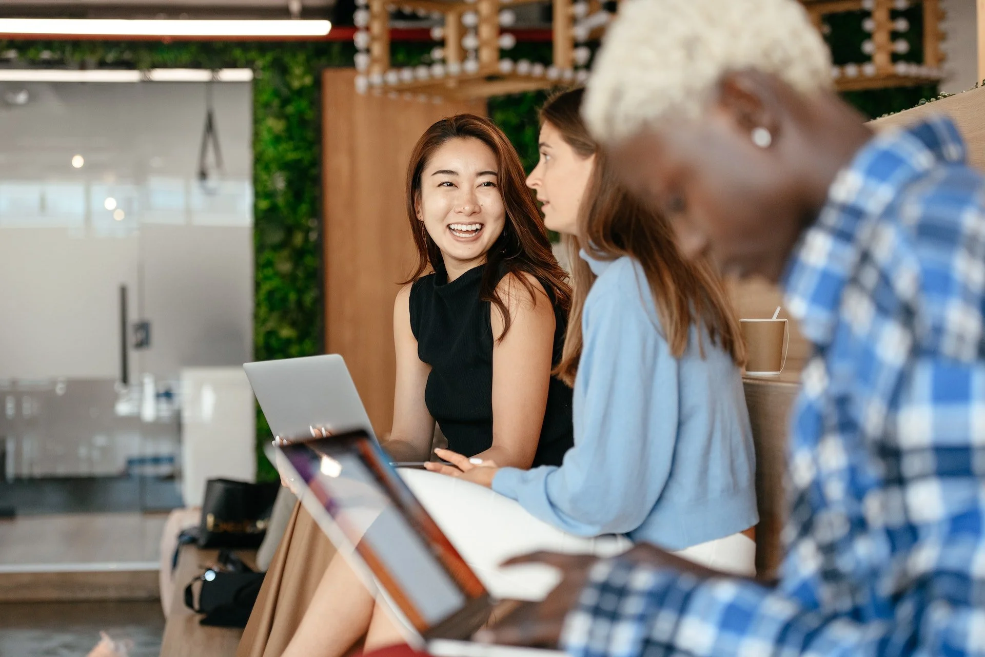 Three women sitting at a table in a modern, green indoor space, engaging in conversation and smiling, with laptops and drinks on the table.