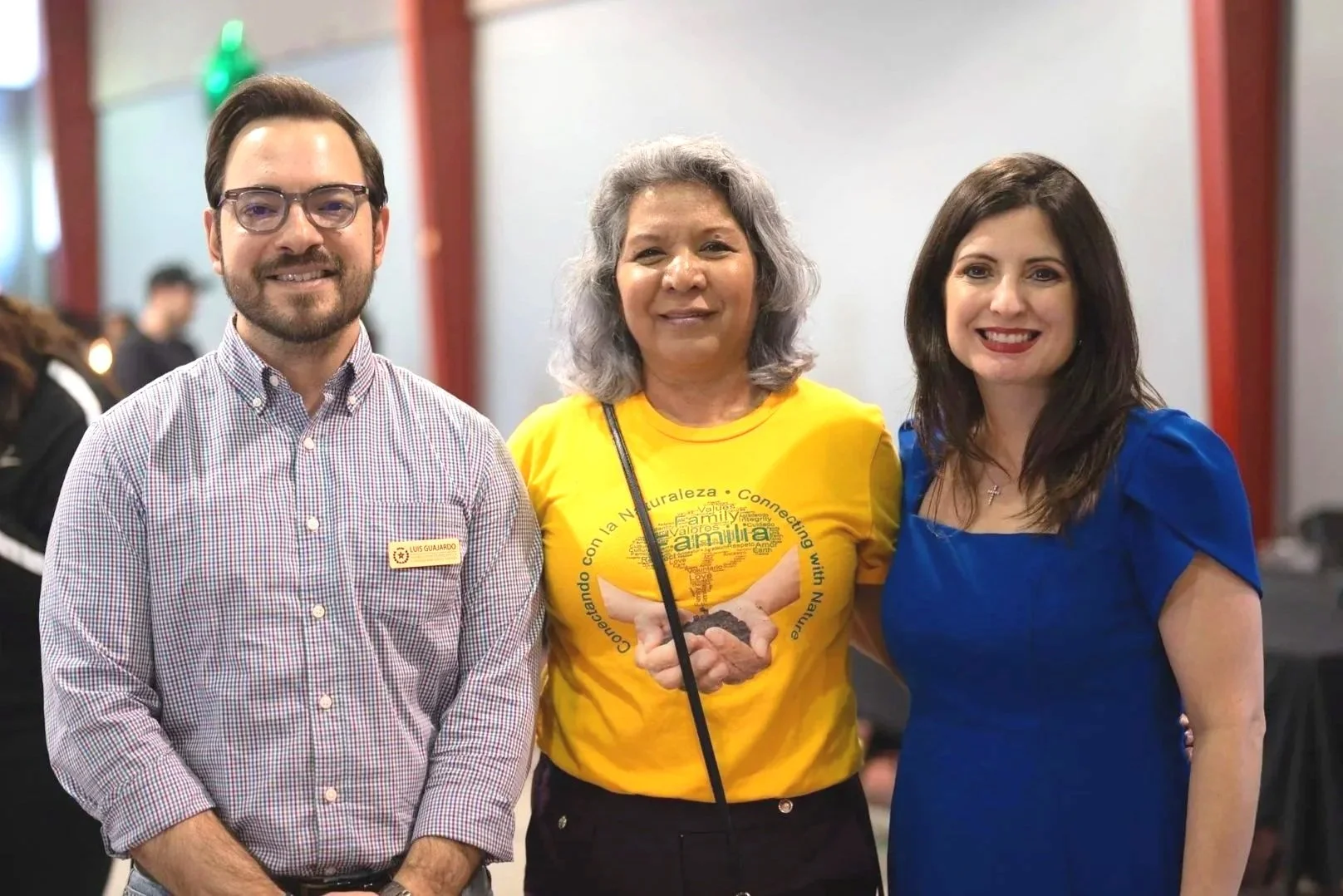 Three people standing close together indoors, smiling at the camera at a Burnett Bayland Park community engagement event.