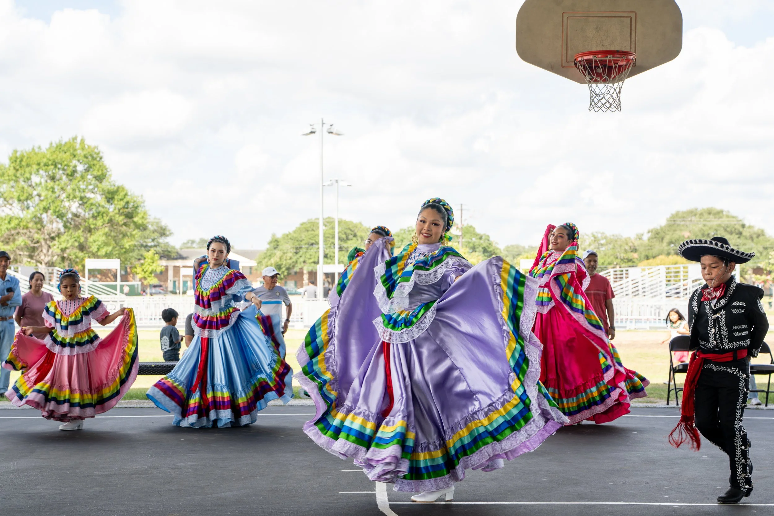 Group of women and children dressed in colorful traditional Mexican dresses performing a folkloric dance outdoors under a basketball hoop with onlookers in the background.