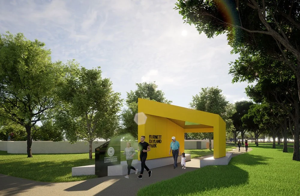 People entering Burnett Bayland Park through a modern yellow entrance structure in a park with green grass and trees, under a partly cloudy, sunlit sky.