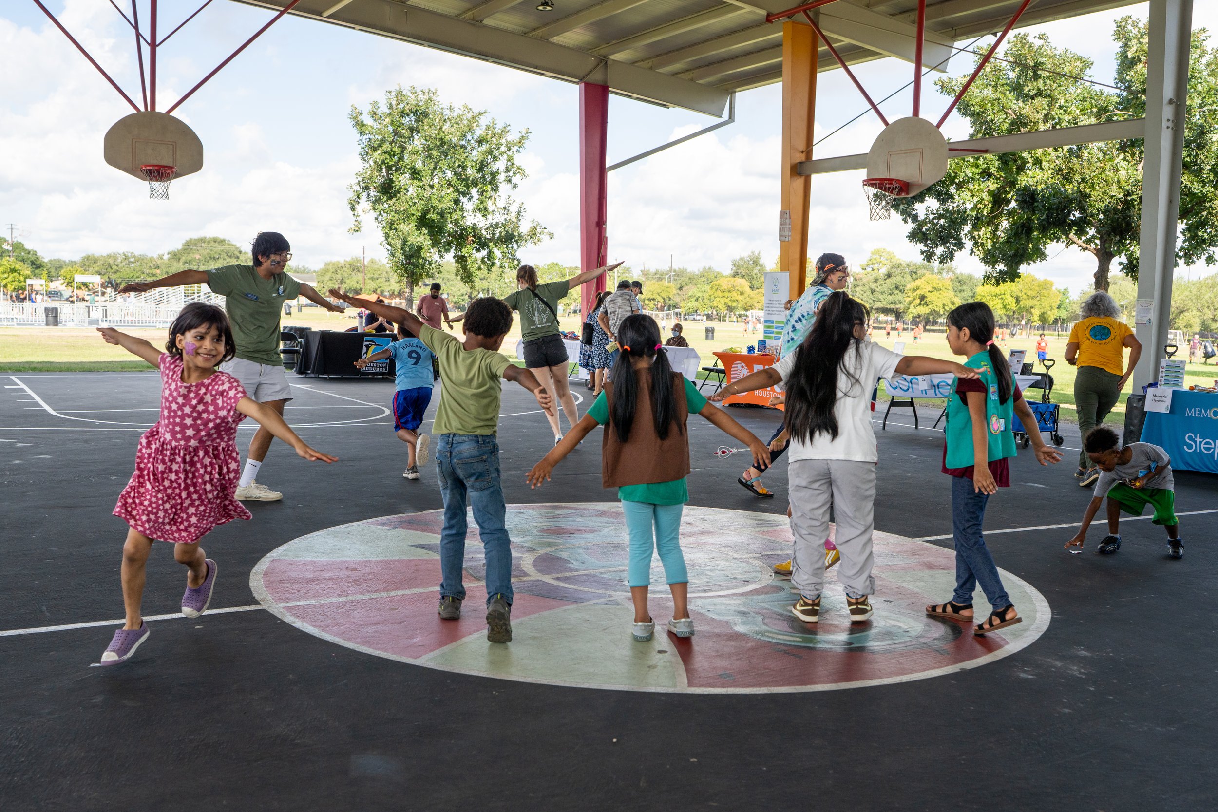 Children and adults dancing and playing at an outdoor event under a covered pavilion with basketball hoops. There are trees and open field in the background.