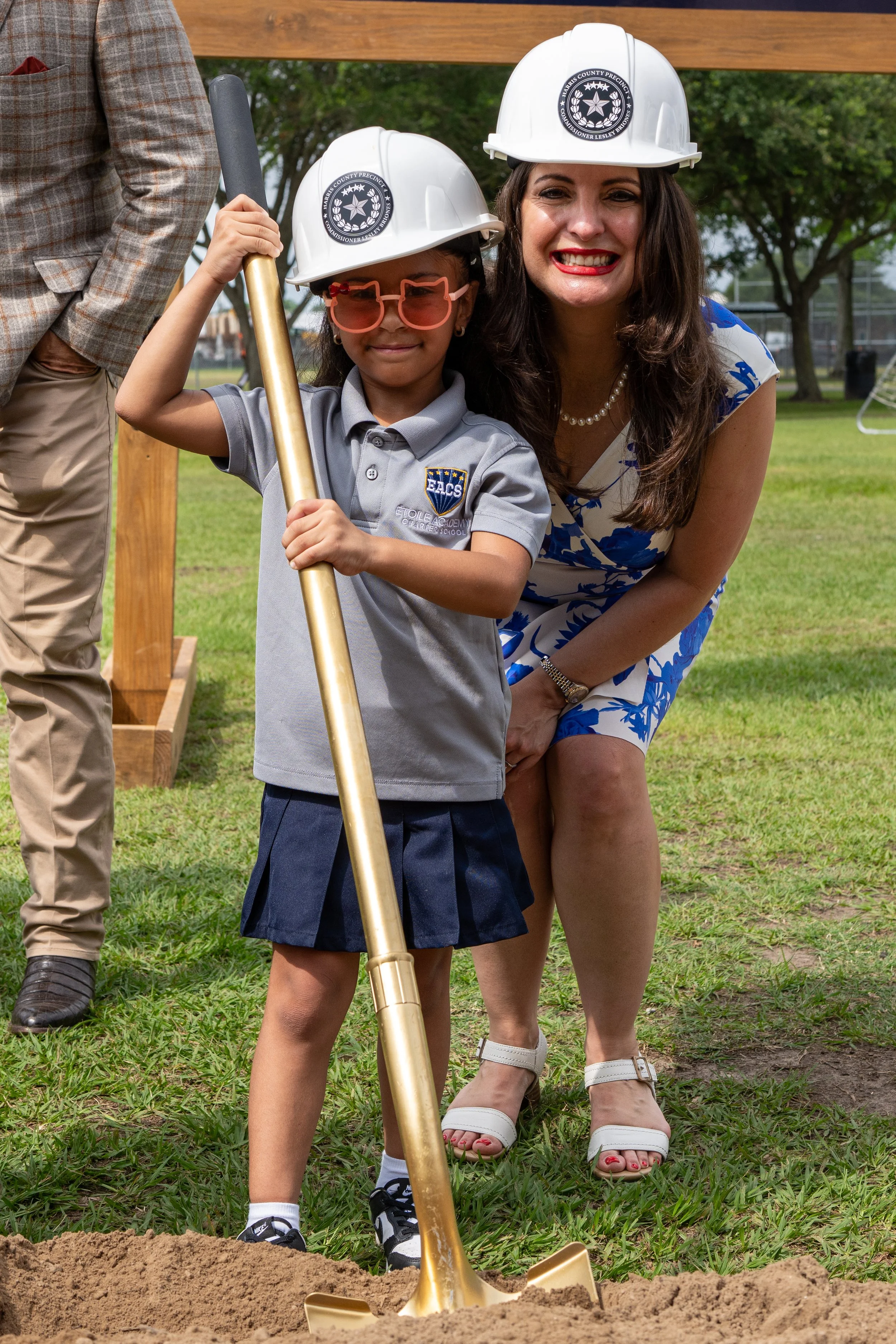 A young girl and a woman, both wearing white construction helmets with a crest and star emblem, are at a groundbreaking ceremony. The girl is holding a golden shovel and wearing pink glasses, a gray school uniform, and black sneakers. The woman is in