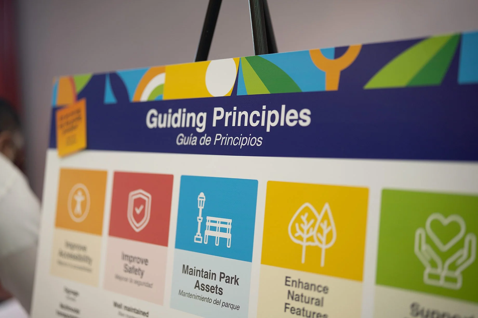 A colorful poster displayed at a Burnett Bayland Park community engagement event titled 'Guiding Principles' in English and Spanish, featuring icons and text about park maintenance, safety, natural features, and sustainability.