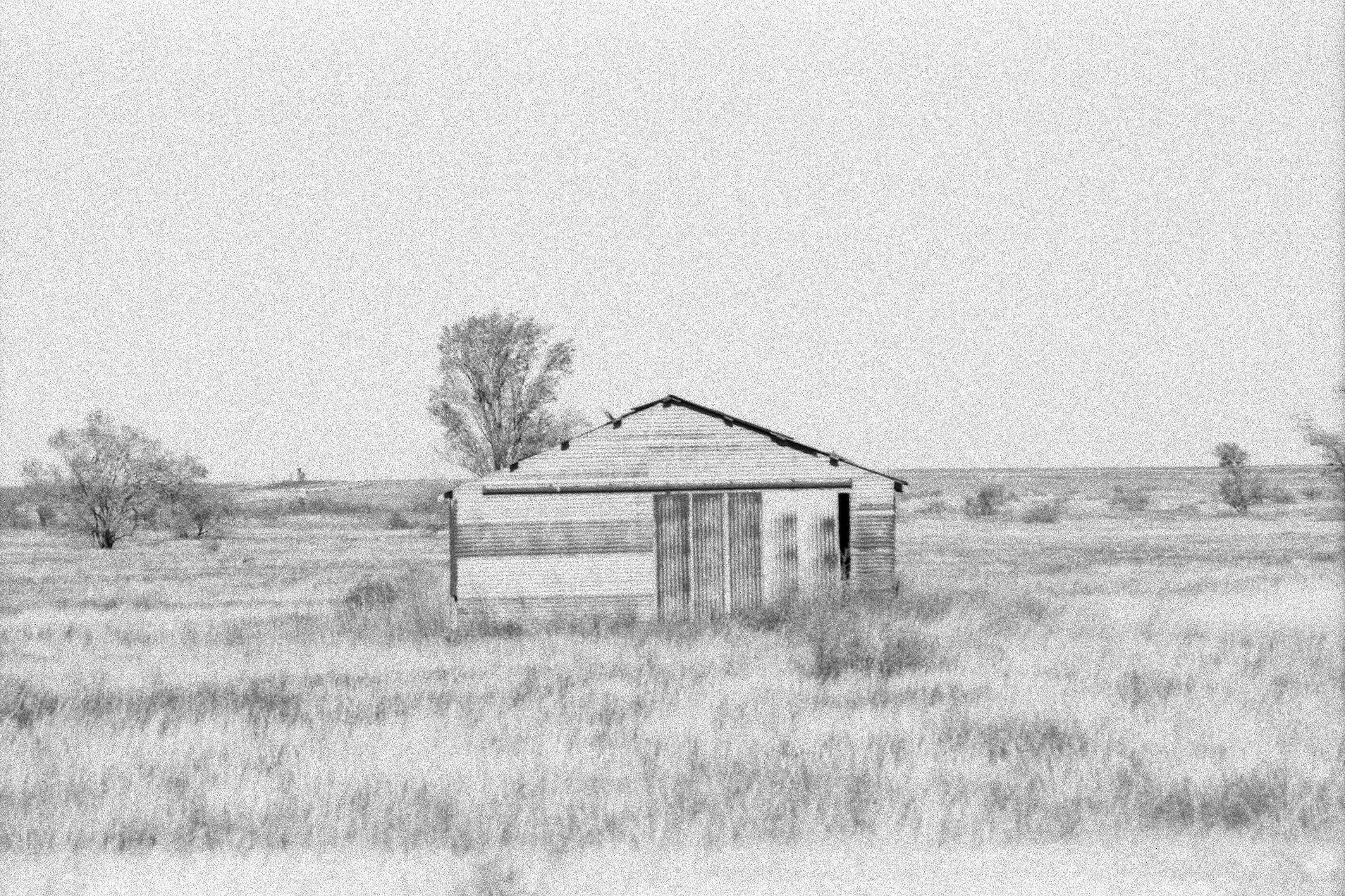 A black and white photograph of a rural landscape featuring a small, old wooden shed with a sloped roof and a door, surrounded by open fields and sparse trees in the background, under a cloudy sky.