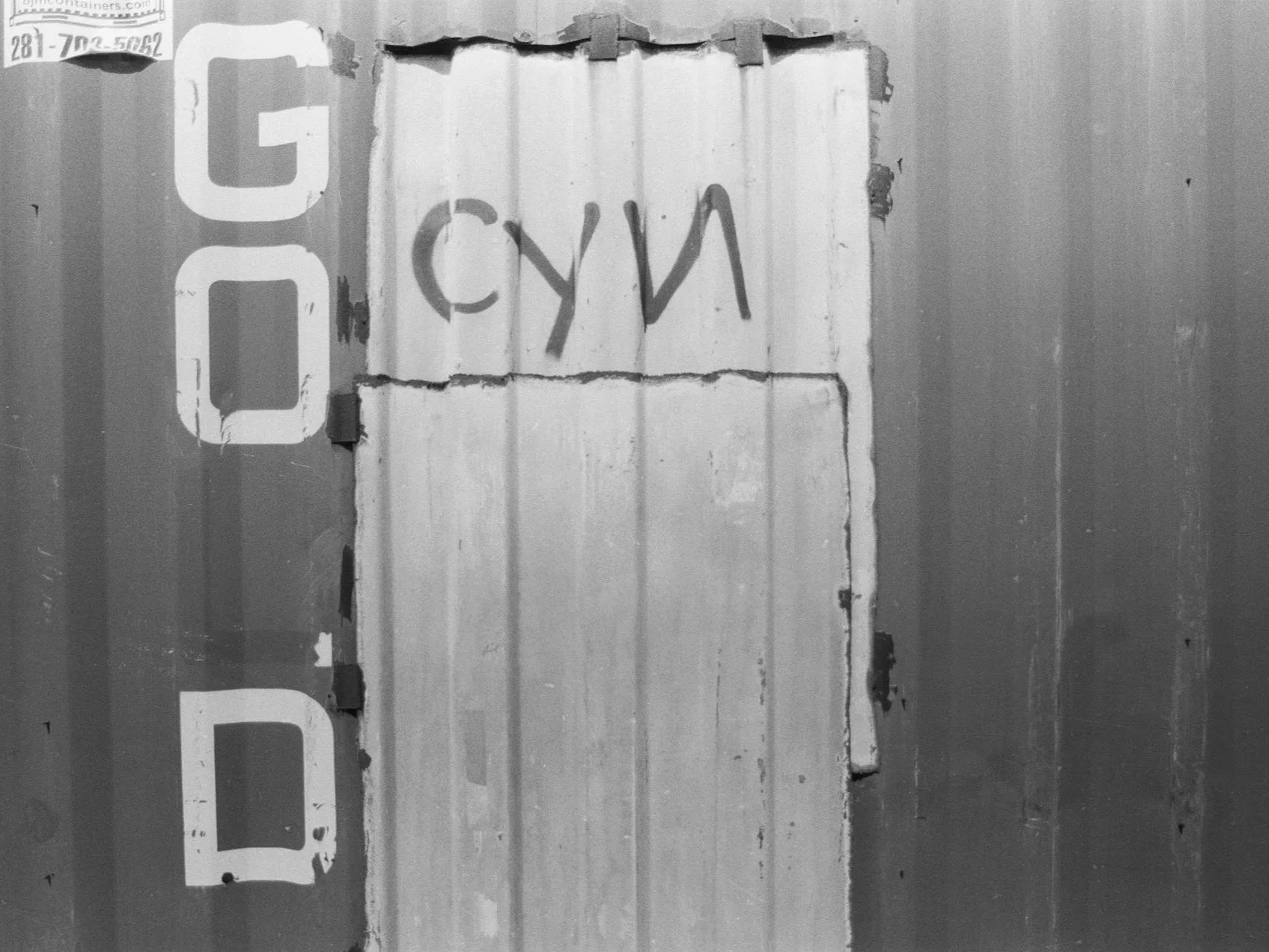 A black and white photo of a weathered wooden door with graffiti that spells out 'CYAN' in spray paint. Part of a vertical sign with large, partially visible letters is seen on the left side of the image.