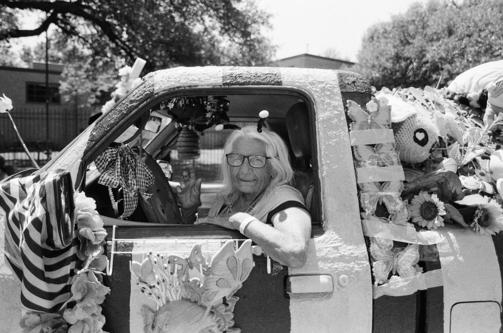 A woman with glasses and long hair sitting in a decorated parade float, smiling, with her hand raised. The float is adorned with flowers, large butterflies, and patterned fabric, and is part of an outdoor event with trees and a building in the backgr