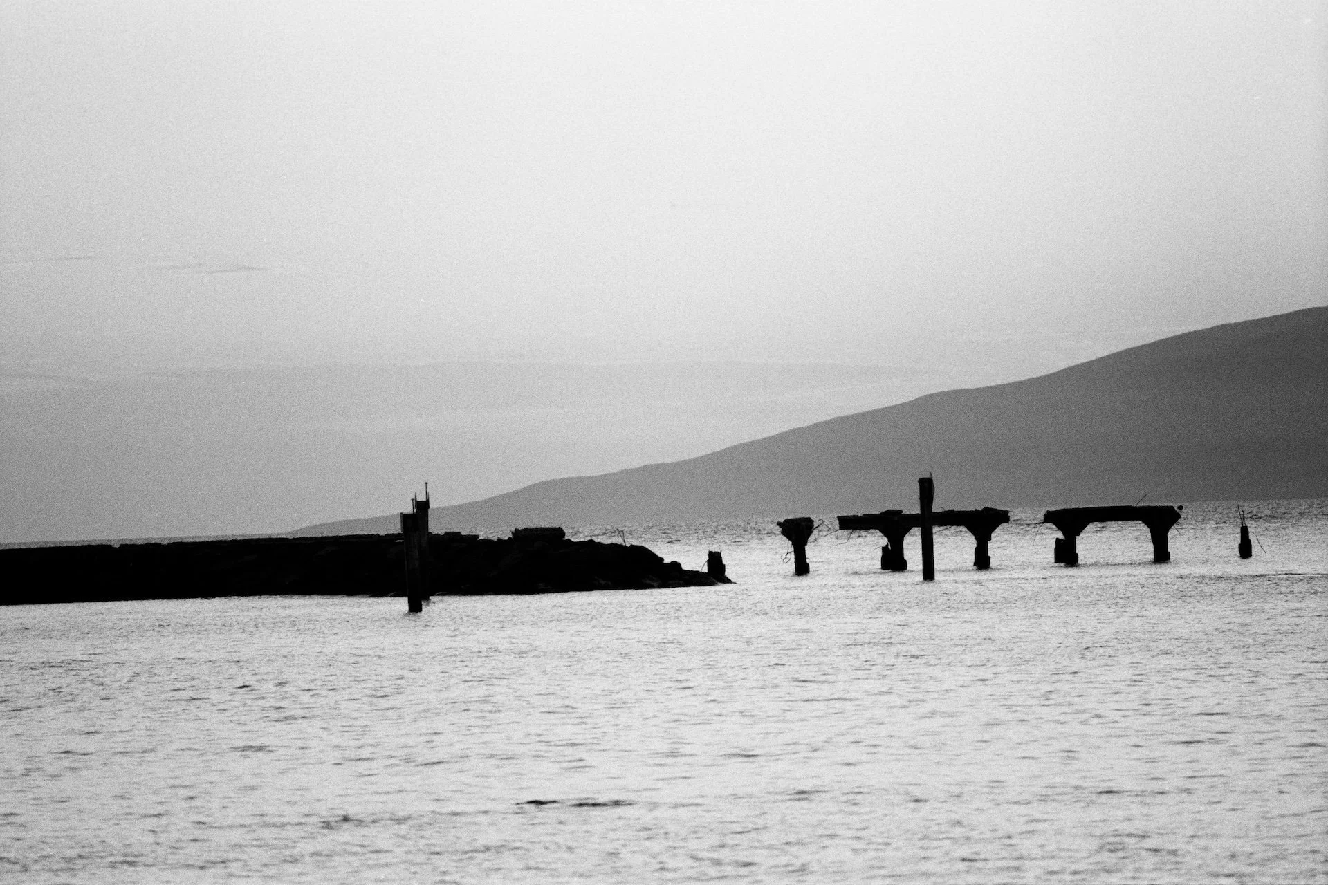 Black and white photo of a calm sea with an old, abandoned pier with broken supports, a rocky shoreline, a small landmass in the distance, and a faint mountain range on the horizon.