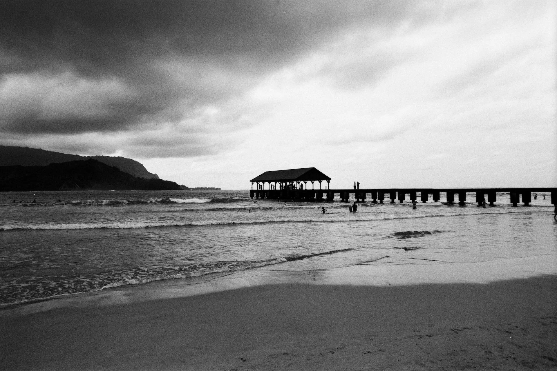 A black and white photo of a beach with waves crashing onto the shore, a pier with a pavilion extends into the water, and a hilly landscape in the background under a cloudy sky.