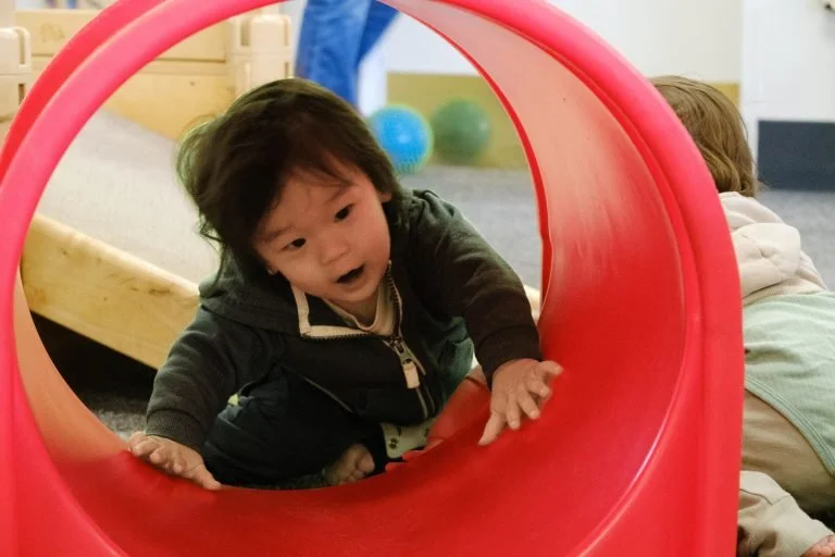 Young child crawling through a red plastic tunnel at a children's play area.