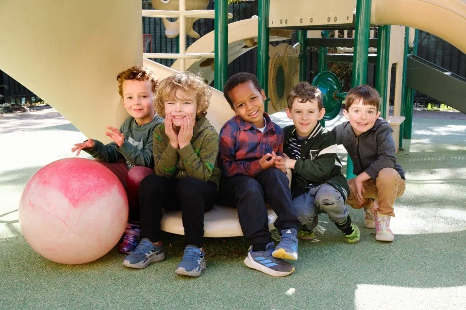 Five young children sitting on a playground slide, smiling and playing with a red ball nearby.
