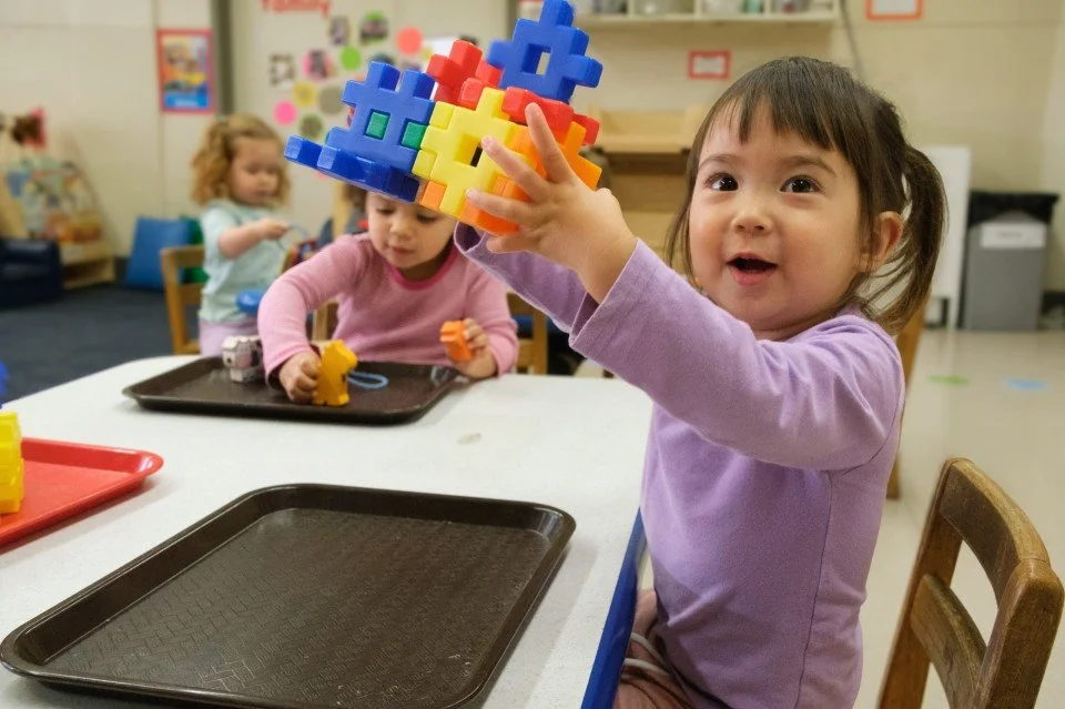 Young girl in a purple shirt holds colorful plastic building blocks at a table in a classroom with other children in the background.