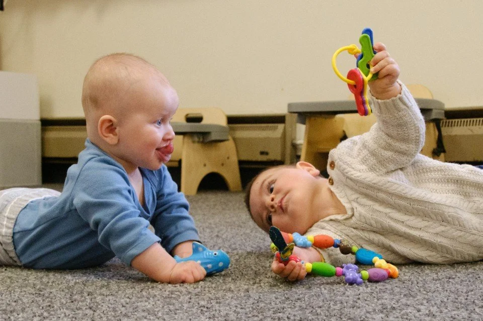 Two young children, a baby and a toddler, lying on the carpeted floor of a playroom. The baby, wearing a blue shirt, is on their stomach and looking at the toddler. The toddler, wearing a white sweater, is lying on their side holding colorful plastic
