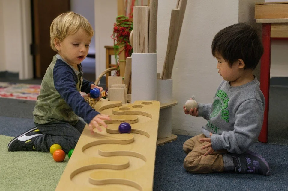 Two young boys playing with wooden balls and a maze toy in a playroom.