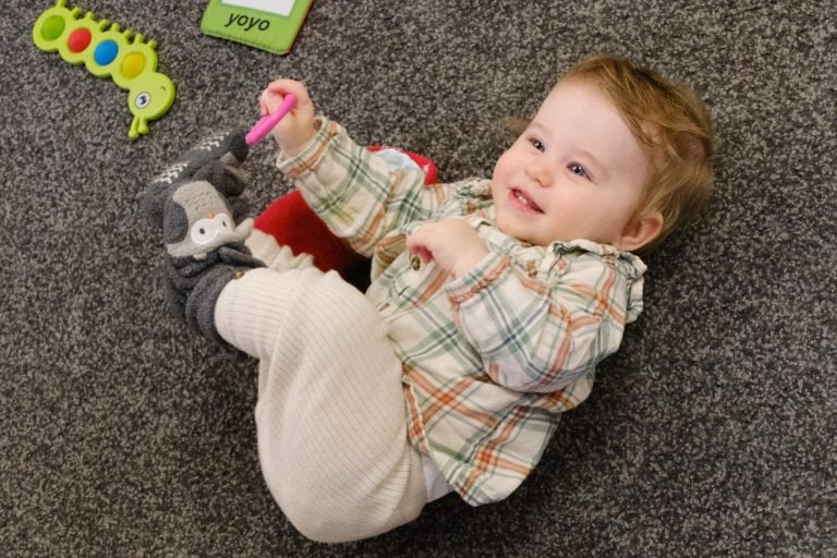 Child lying on a carpeted floor, playing with toys, smiling, wearing beige pants, a plaid shirt, and socks with animal slippers.