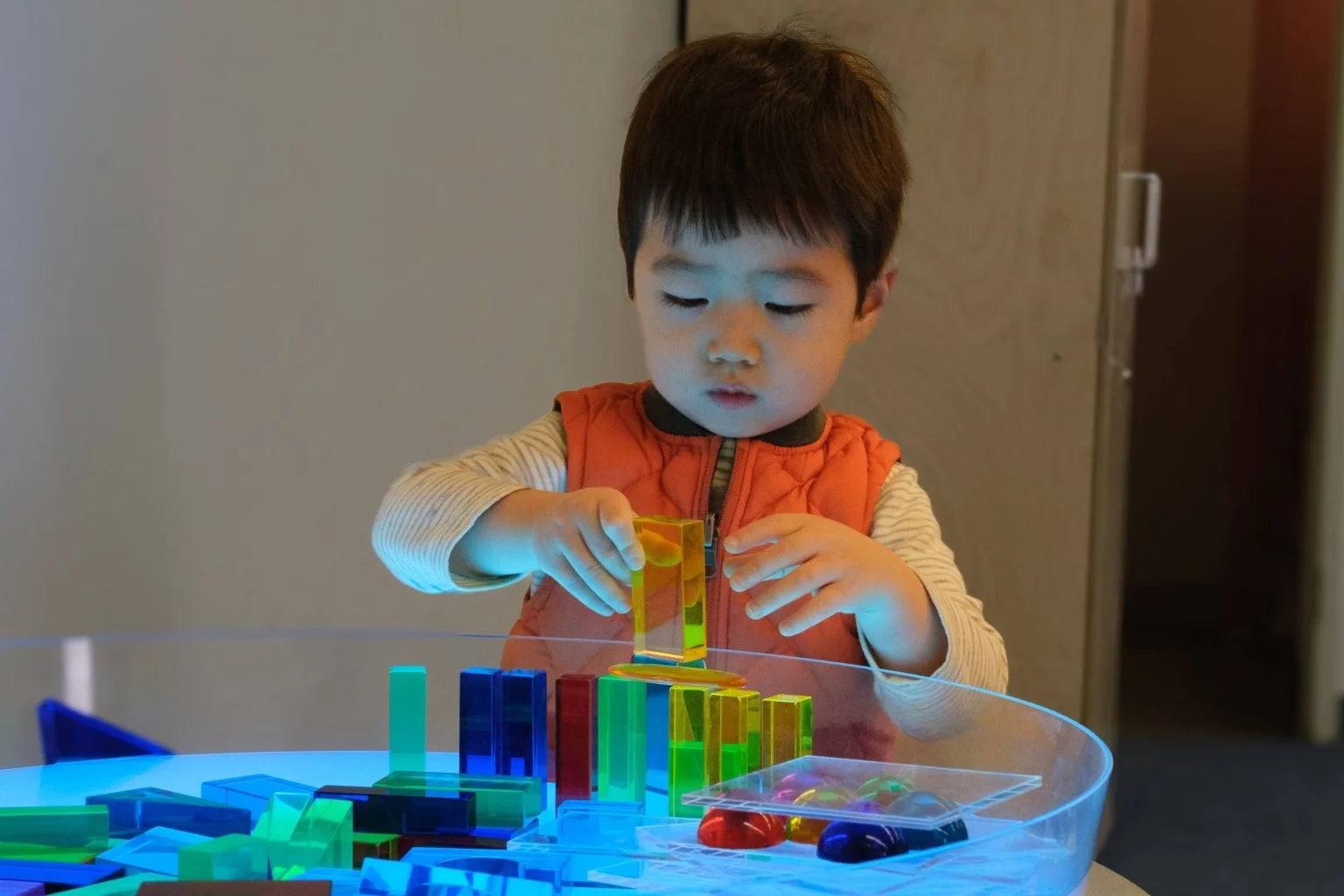 A young boy playing with colorful magnetic tiles on a table, focusing on stacking a yellow tile.