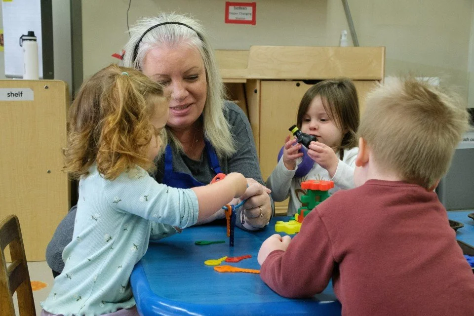 A woman and three young children playing with colorful plastic toys on a blue table in a classroom setting.