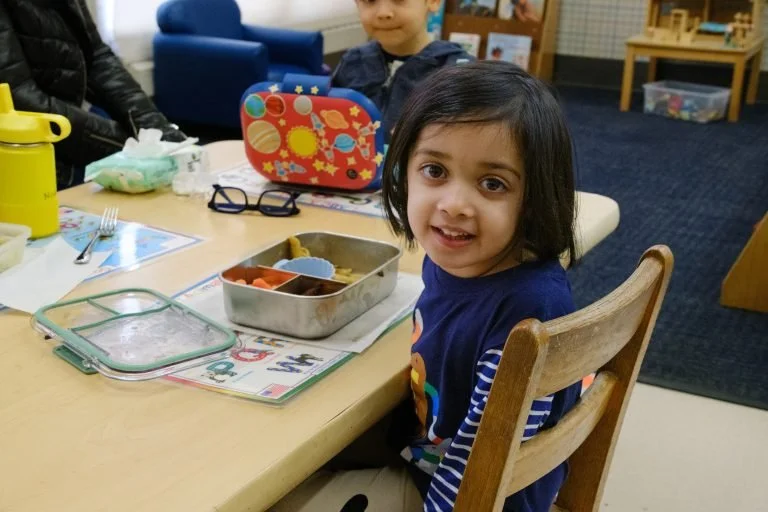 A young girl sitting at a dining table with a lunchbox, glass of water, and utensils, smiling at the camera in a classroom or daycare setting.