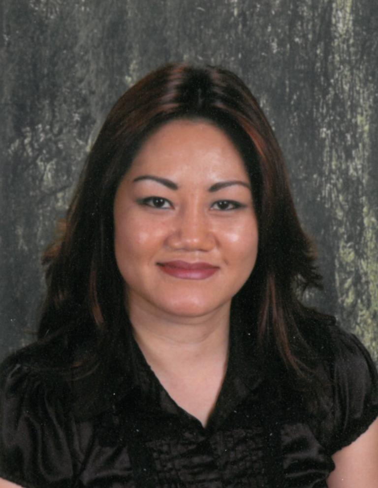 A woman with shoulder-length dark hair, smiling, wearing a black blouse, with a blurred nature background.