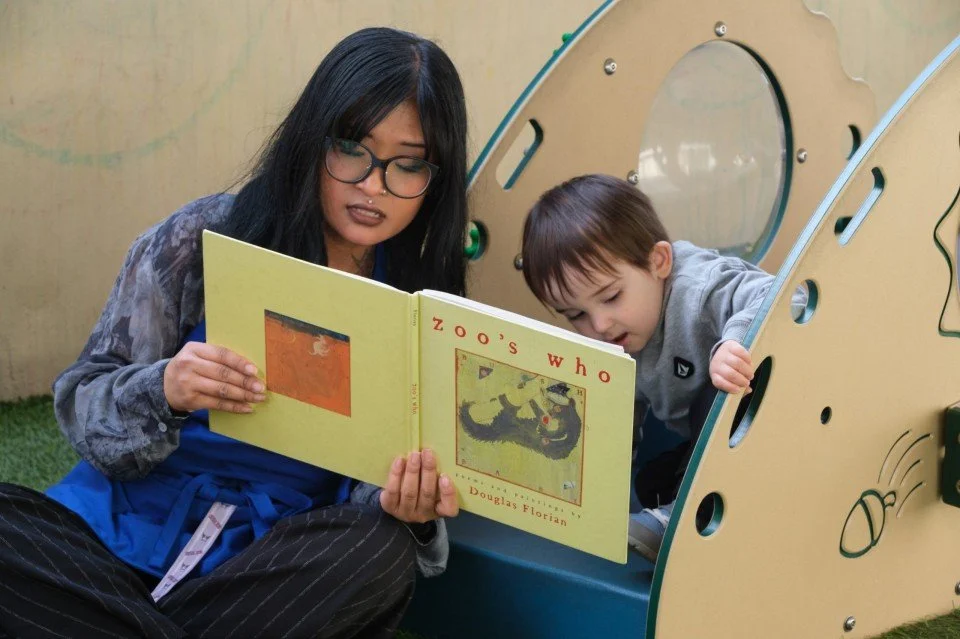 A woman reading a children's book titled 'Zoo's Who' to a young child next to a playground slide.