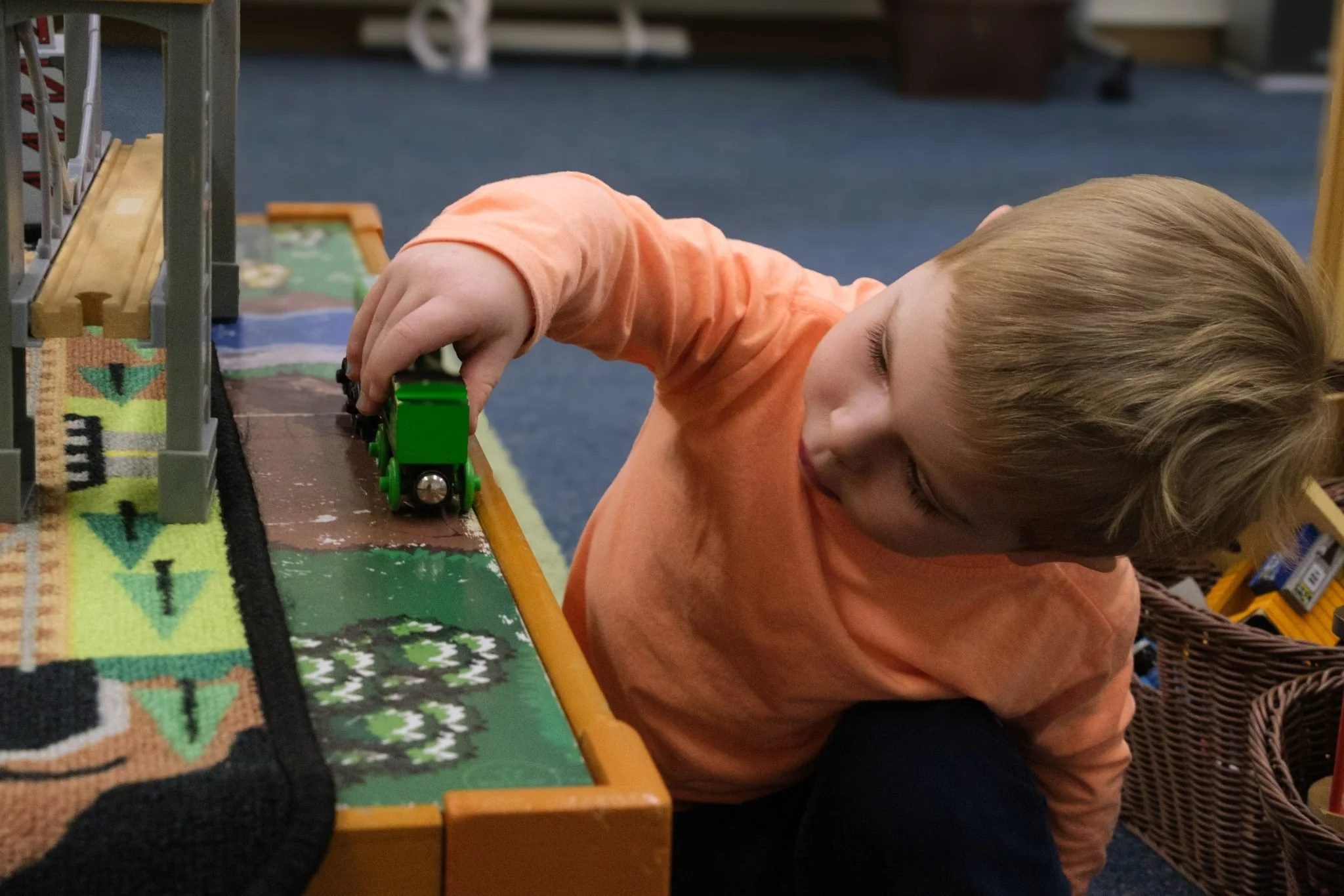 A young boy with blonde hair laying on a table, playing with a green toy train. The table has a colorful textured surface resembling a miniature landscape or game.