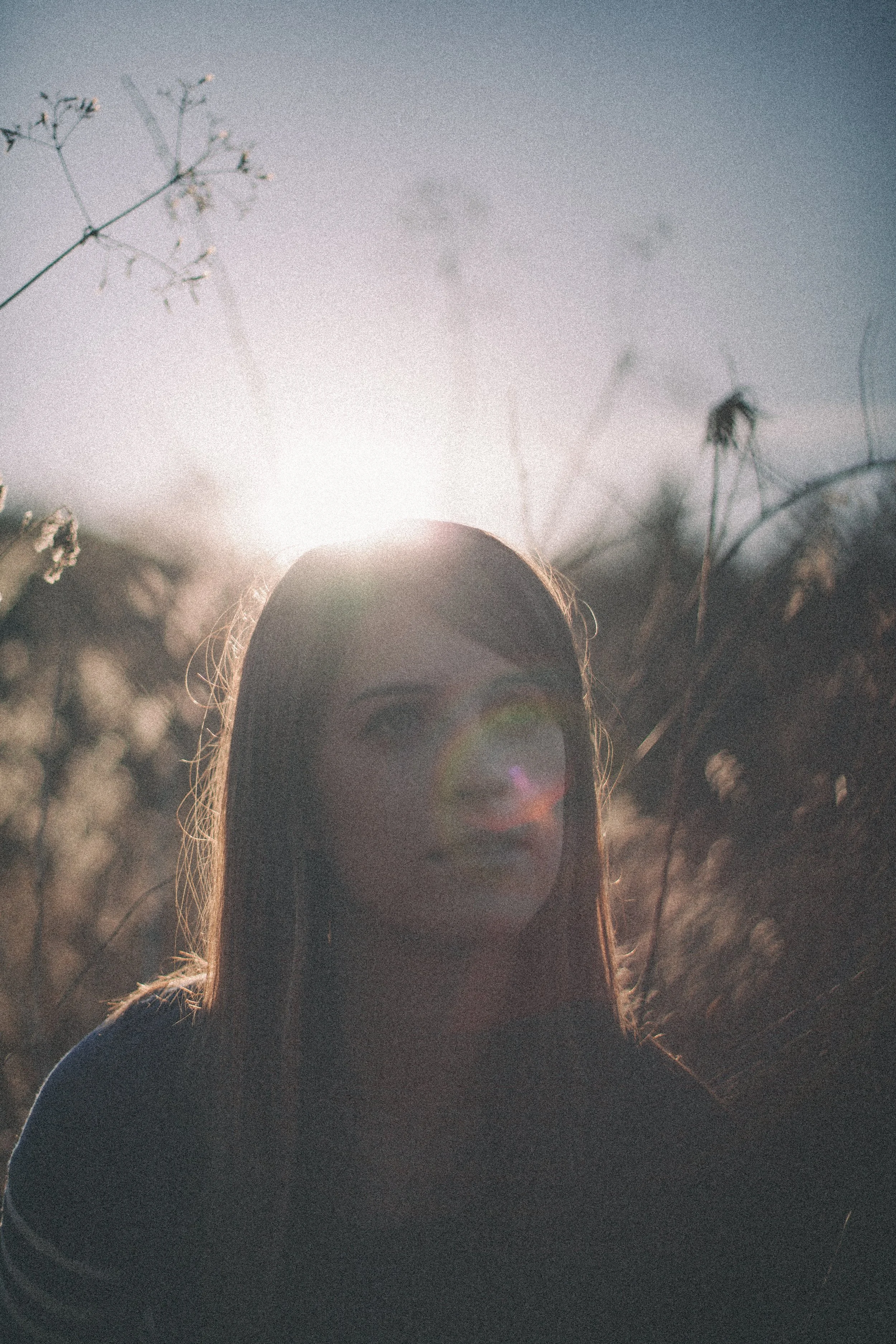 A person in a dark hat standing outdoors during sunset with sunlight shining in from behind, creating a silhouette effect, with some plants and trees in the background.