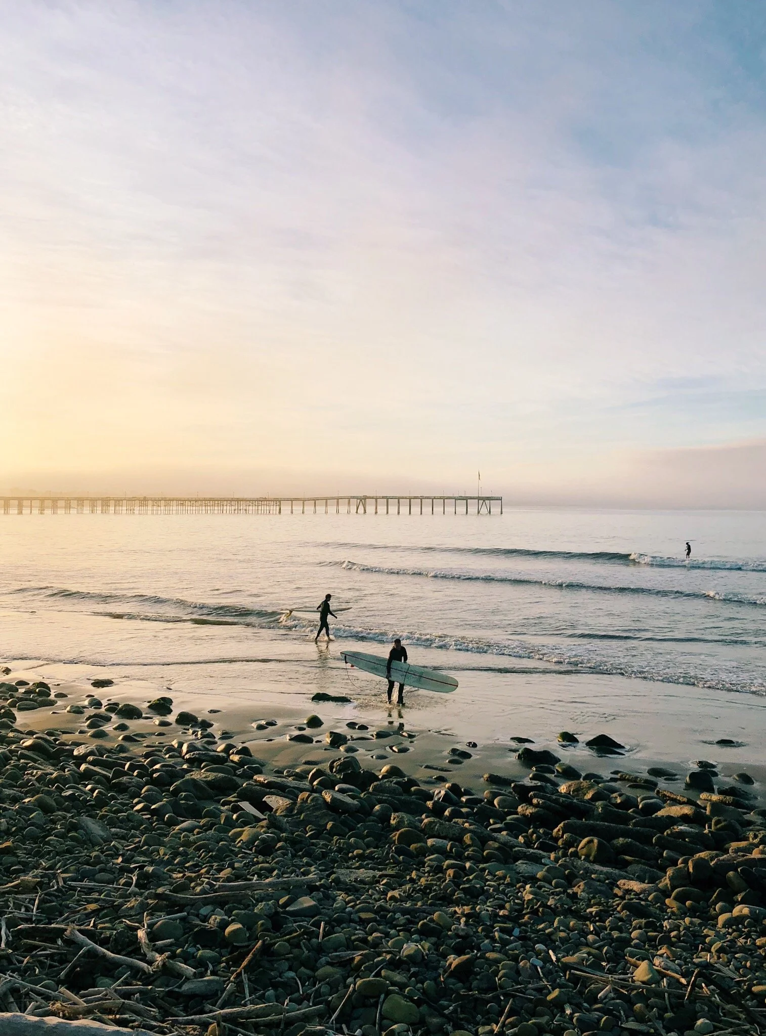 A scenic view of a beach during sunset, with a wooden pier extending into the calm ocean and a person holding a surfboard near the water's edge.